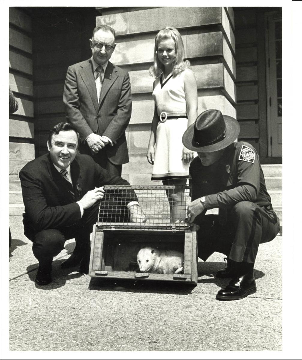 Black and white photograph of a white man in a suit and glasses, a white woman in a dress, a white man in a suit kneeling, and a white man in a police uniform kneeling around a metal cage with Slow Poke the Possum inside. They are standing in the sunshine outside of the State Capitol building.