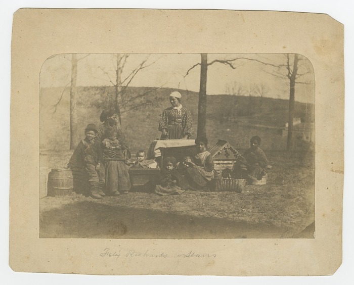 An albumen print on paper with a purple, reddish-brown hue depicting two adult women and seven children pictured.  The group is posed outside in front of bare trees, one woman is posed as if ironing. Baskets and a dog or doll house are placed around the group. The women and their children were enslaved at the time this photograph was taken on a plantation just west of Alexandria, Virginia, that belonged to Felix Richards.