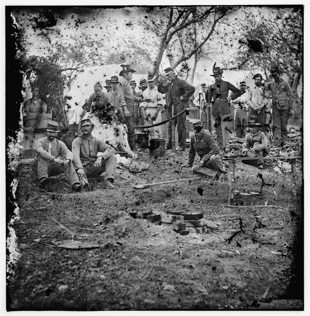 Civil War soldiers gathered around a fire, preparing food. At an encampment, a pot hangs from a frame made of sticks.