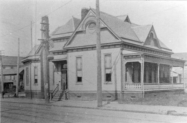 Photograph of the Durham County Public Library, 1900. A small wooden building with a porch and chimney. It is white.