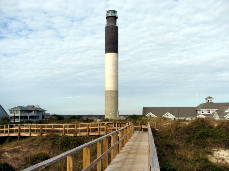 A tall lighthouse stands over homes and sand dunes. It is tiered with gray, white, then black brick. The sky is cloudy. 