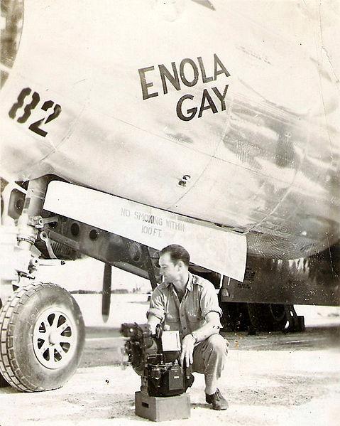Enola Gay bombardier Thomas Ferebee with the Norden Bombsight on Tinian after the dropping of Little Boy. A pilot sits beside an aircraft labeled "Enola Gay."