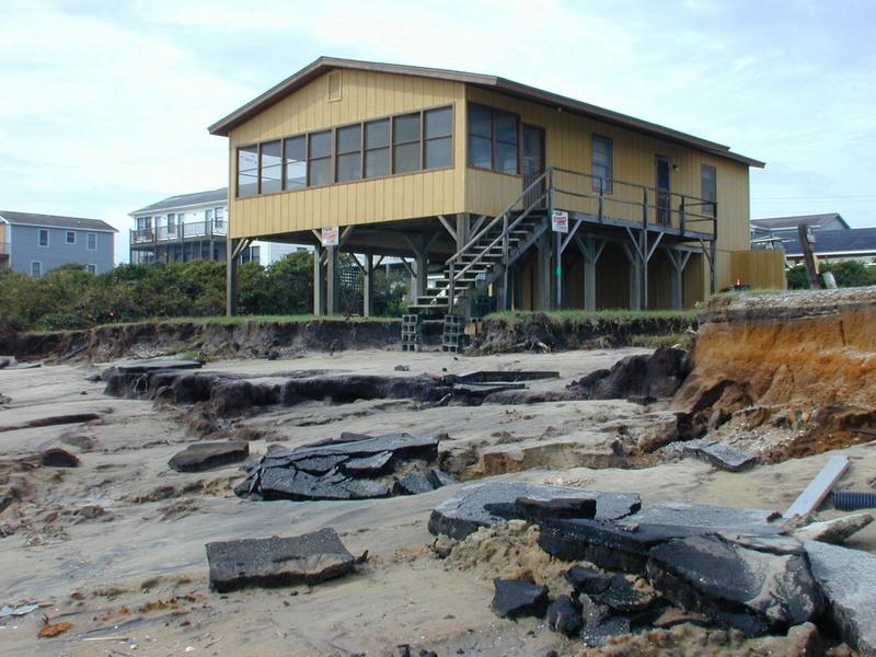 Yellow house on a ruined beach in Oak Island after Floyd. Broken pieces of the road litter the beach. There is a blue sky in the background. 