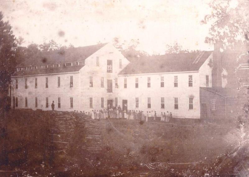 Alamance Cotton Mill. It is a sepia print. There are people in front of the mill, which is a large building attached to a smaller building. There are trees around. 
