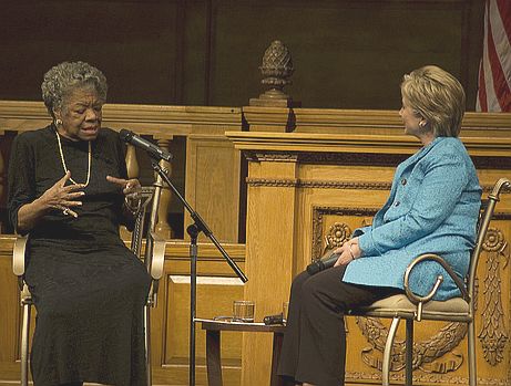 Maya, seated left, speaks into a microphone. She is wearing a black dress with a long gold necklace. Her hair is short and wavy and styled back from her face. Hillary Clinton is seated to Maya's right, wearing a sky blue blazer and listening intently. 