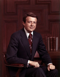Photograph of James Eubert Holshouser, Jr. sitting in front of a wood paneled wall and books.
