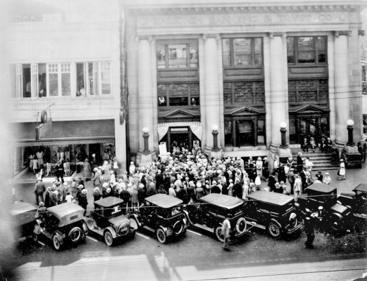 People gather outside a building. There are also cars with them. Black and white photo.