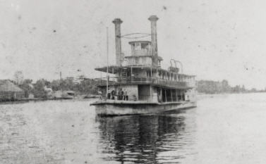 Black and white photo of a boat on water close to the shore. The boat has two large chimney-like towers and three levels.