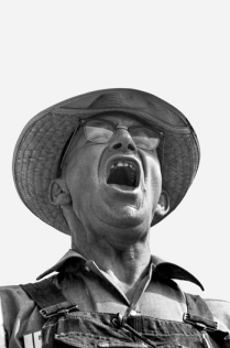 Black and white photograph of a man with his mouth wide-open. He appears to be yelling or singing. He is wearing overalls, glasses, and a wide-brimmed hat.
