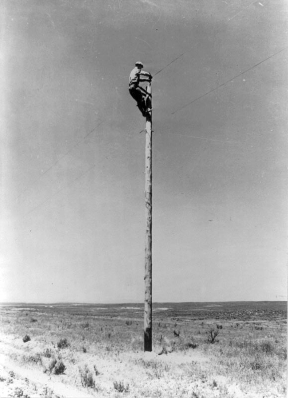 An electrical lineman running lines on top of a tall post. 