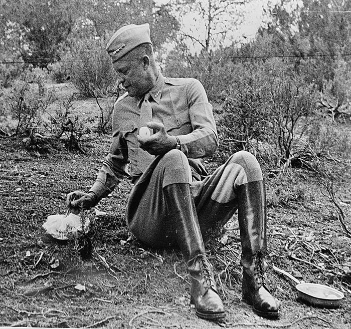 Black and white photo. Eisenhower eating from a ration. He is in his military uniform and he is surrounded by shrubbery. 