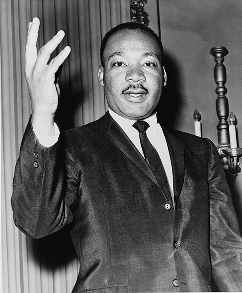 Martin Luther King, Jr. He is posed in a suit with his hand raised, as if giving a sermon. He has a thin moustache and short hair. 