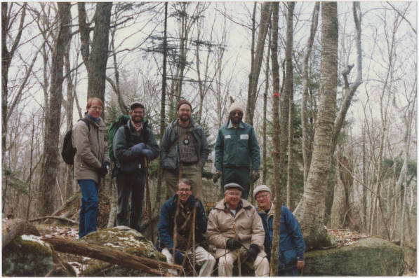 Group of 7 hikers in a wooded area. They are dressed warmly and are all smiling. 