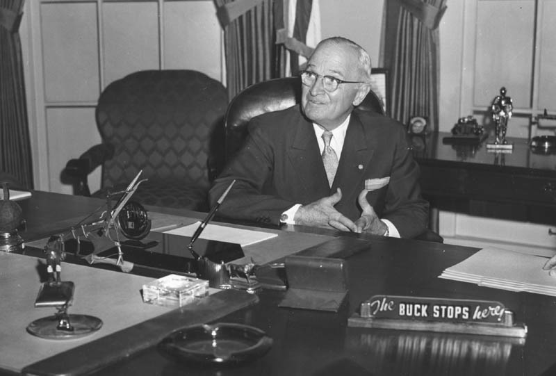 Harry Truman in his presidential office. He is wearing glasses, a suite, and is balding.