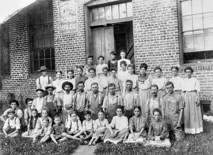 Employees at a textile mill. Most are young and some are children. They look sad. The building is brick.