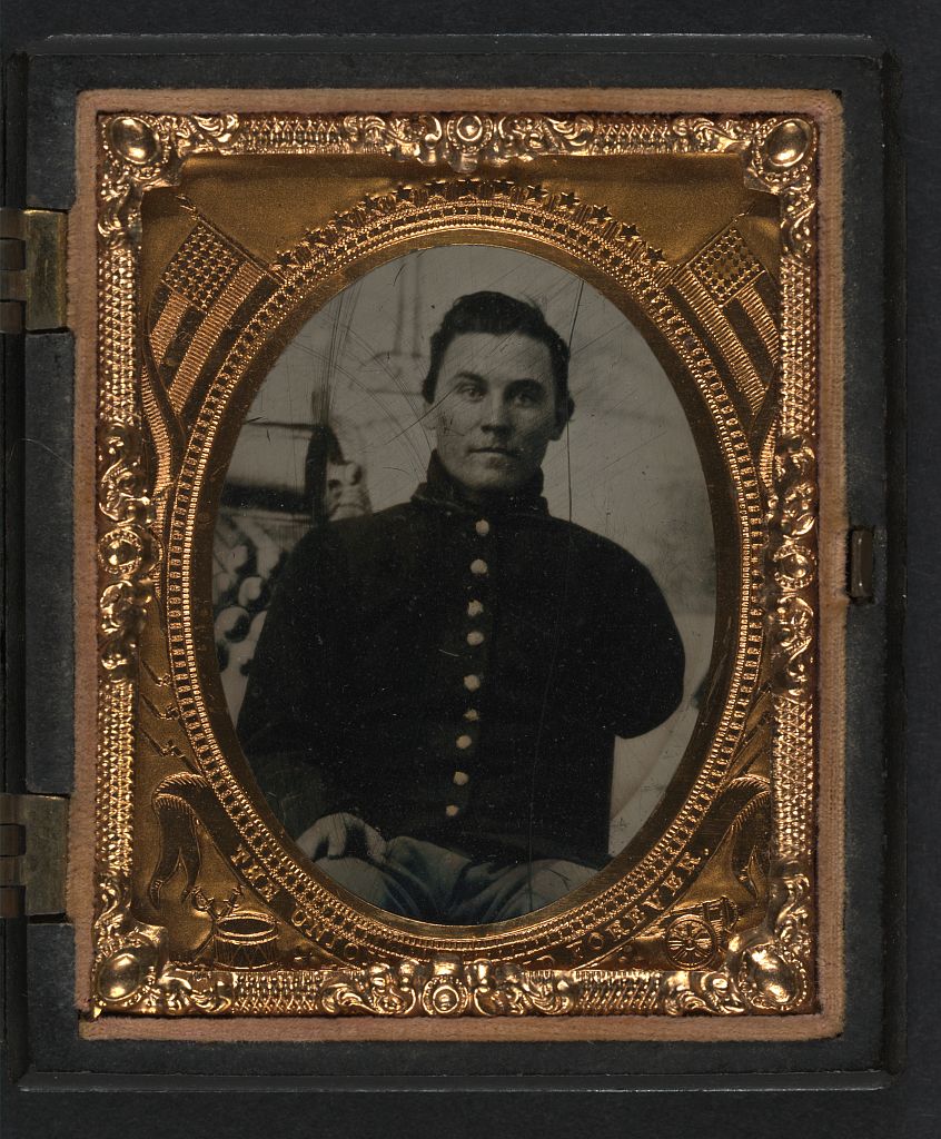 Unidentified soldier with amputated arm in Union uniform in front of painted backdrop showing cannon and cannonballs