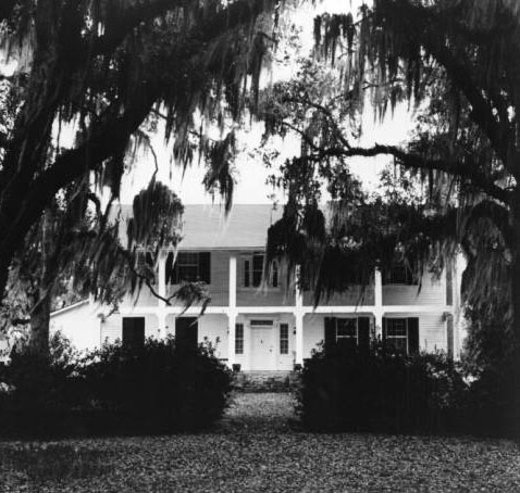Highland plantation It is a large two story house with porches on both levels. There are trees covered in Spanish Moss in the foreground. 