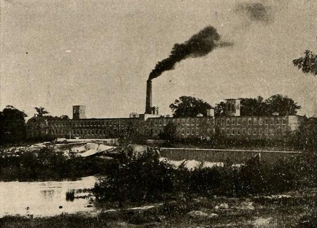 Rocky Mount Mills. A large factory complex sits in the background with an active smokestack. Some small plants are in the foreground.